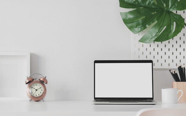 A styled photo of a desk with a clock, a laptop and a leaf